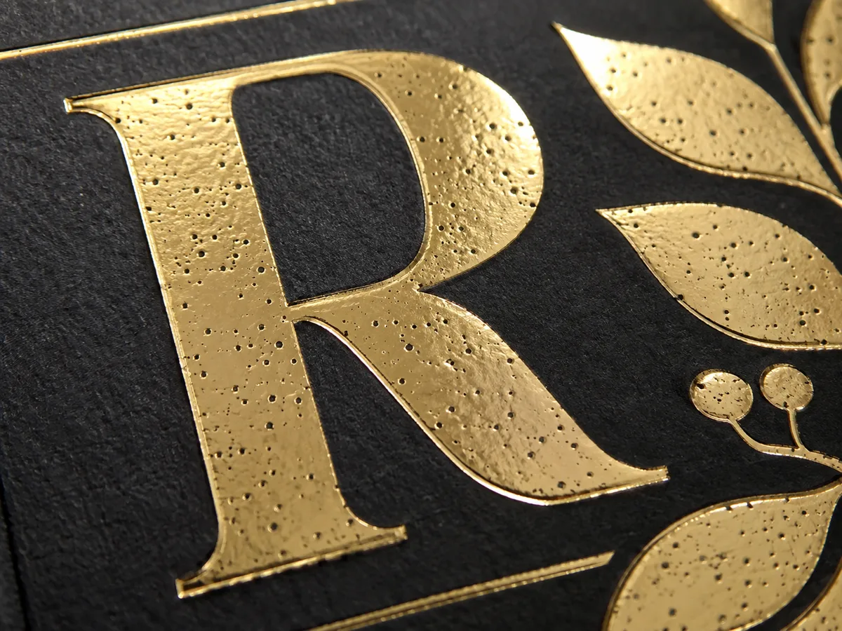 Macro close-up of a gold cold foil monogram letter on black stock showing scattered pinholes across the metallic surface